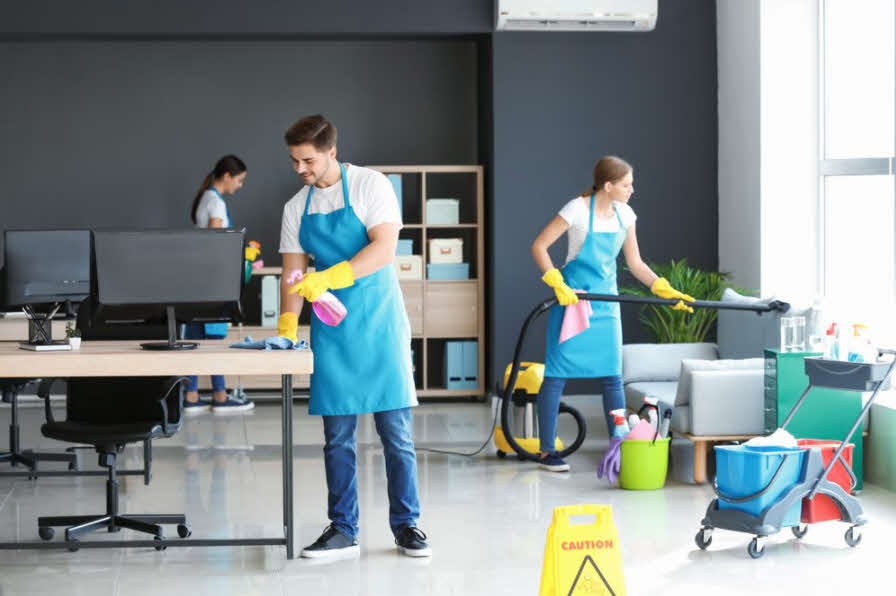 Three people cleaning a computer table and sofa in a commercial space